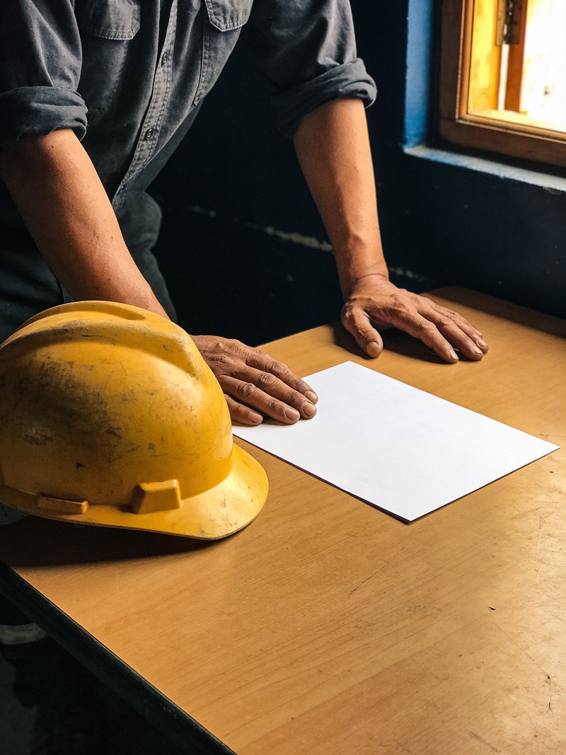 Manos de un trabajador apoyadas sobre una mesa de madera junto a un casco de seguridad amarillo y una hoja en blanco